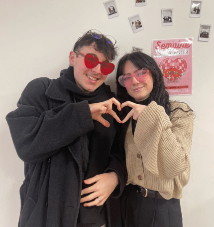 Un jeune homme et une jeune femme posent ensemble en formant un cœur avec leurs mains. Ils portent des lunettes en forme de cœur, rouges et roses, et affichent de larges sourires. En arrière-plan, on aperçoit un mur décoré de photos et une affiche annonçant la "Semaine Saint-Valentin". L’ambiance est chaleureuse et festive. 💕😊